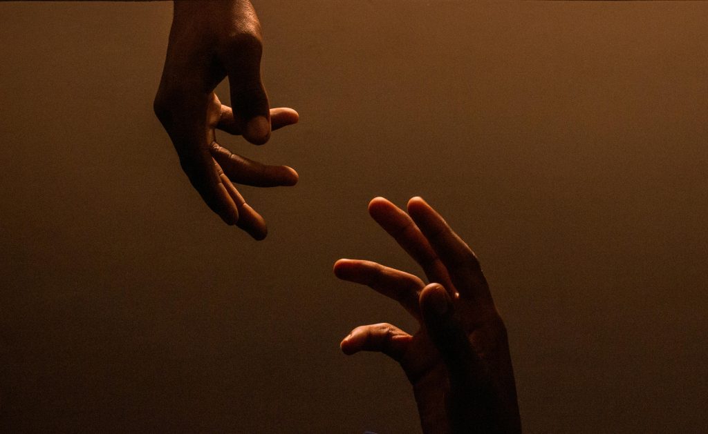 one hand reaching down; another reaching up; space in between (not touching); brown backlit background